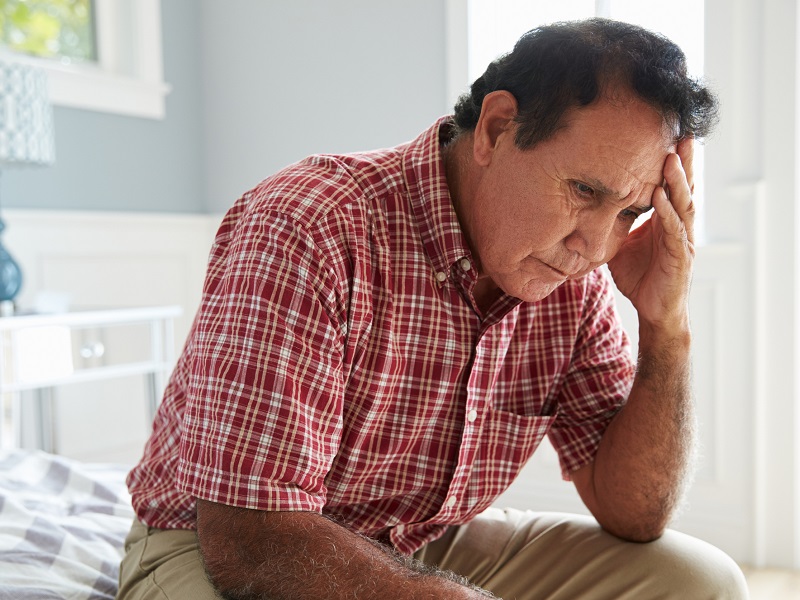 Older man sitting at the edge of a bed, looking confused